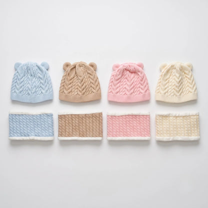 Four cable-knit baby hats with ear details in light blue, tan, pink, and cream, displayed above matching knit neck warmers on a light gray background.