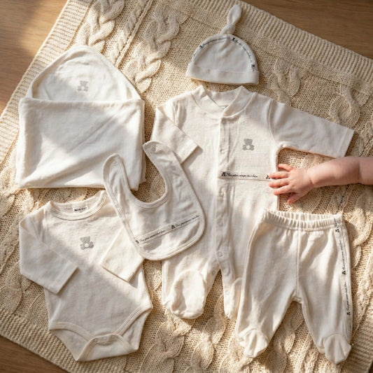 Collection of baby clothes including a onesie, pants, bib, and hats on a textured surface.