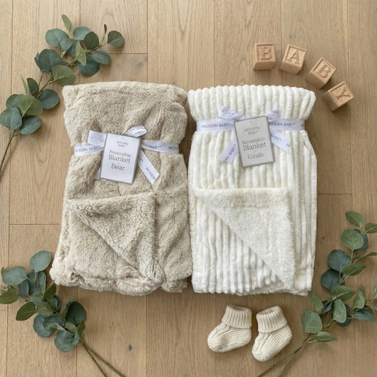 Two baby blankets, a pair of socks, and wooden blocks on a wooden surface with eucalyptus leaves.