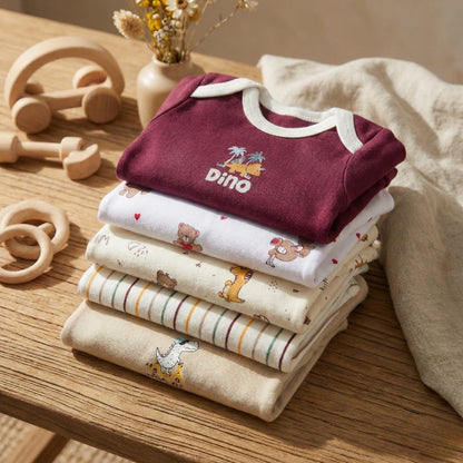 Stack of baby clothes with a maroon onesie featuring 'Dino' on a wooden surface with toys and a towel.