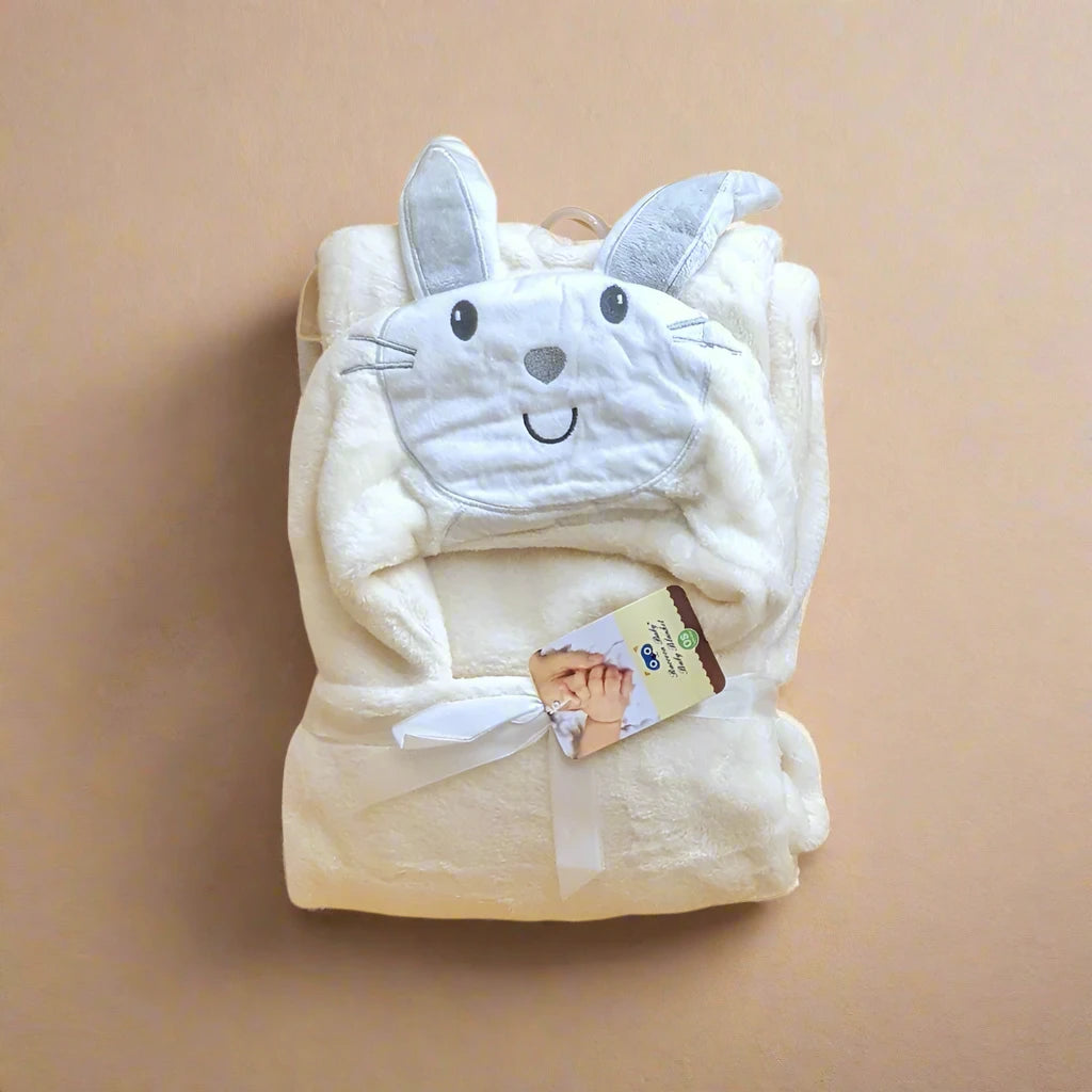 A soft, cream-colored plush Character Hooded Blanket/Wrapping Sheet – Winter (100 x 70 cm) from Alaya Junior is folded neatly. This versatile baby essential features an attached hood with adorable rabbit ears and an embroidered rabbit face. A white ribbon secures the blanket, complemented by a small tag featuring a baby's hand image and product information.