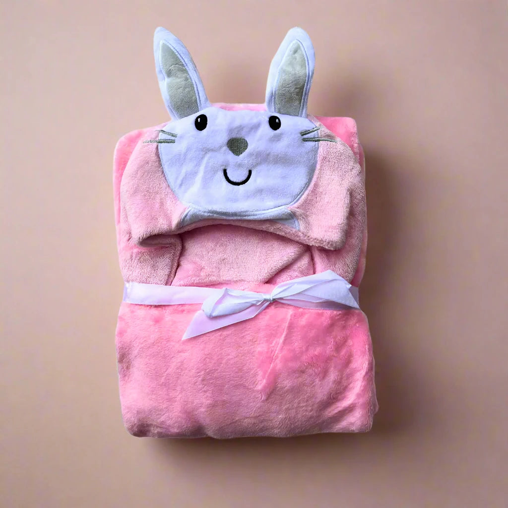 A neatly folded Character Hooded Blanket/Wrapping Sheet – Winter (100 x 70 cm) by Alaya Junior, made from plush pink material, featuring a hood designed to resemble a bunny's face complete with ears and a smiling expression. The blanket is secured with a white ribbon tied in a bow and set against a plain beige background, showcasing one of the cutest character hood designs.