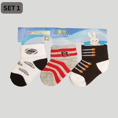 A pack of three baby socks, perfect for newborns, labeled "2023 Trio Pack: Children's Cartoon Cotton Socks for All Seasons" by Alaya Junior. The designs include black and gray fish-like shapes, red, white, and gray stripes with a letter "F," and orange and gray vehicle motifs on a black background. Ideal for infant clothing needs in Pakistan.