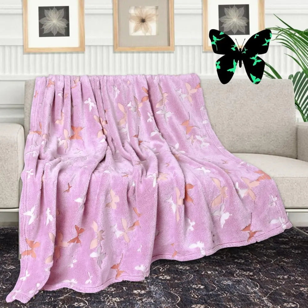 A cozy, purple Glow in the Dark Blanket from Alaya Junior, featuring butterfly and star patterns, is draped over a beige sofa in a snug living room. A decorative framed butterfly print hangs on the wall behind the sofa, enhancing the room's nature-inspired decor.