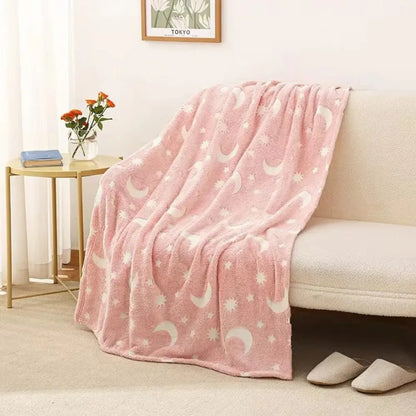 An Alaya Junior Glow in the Dark Blanket, featuring whimsical butterfly and star patterns, is draped over a light-colored sofa. Next to the sofa, a side table with a book and a vase of red and white flowers enhances the homey ambiance. A pair of slippers is placed neatly on the carpeted floor nearby.