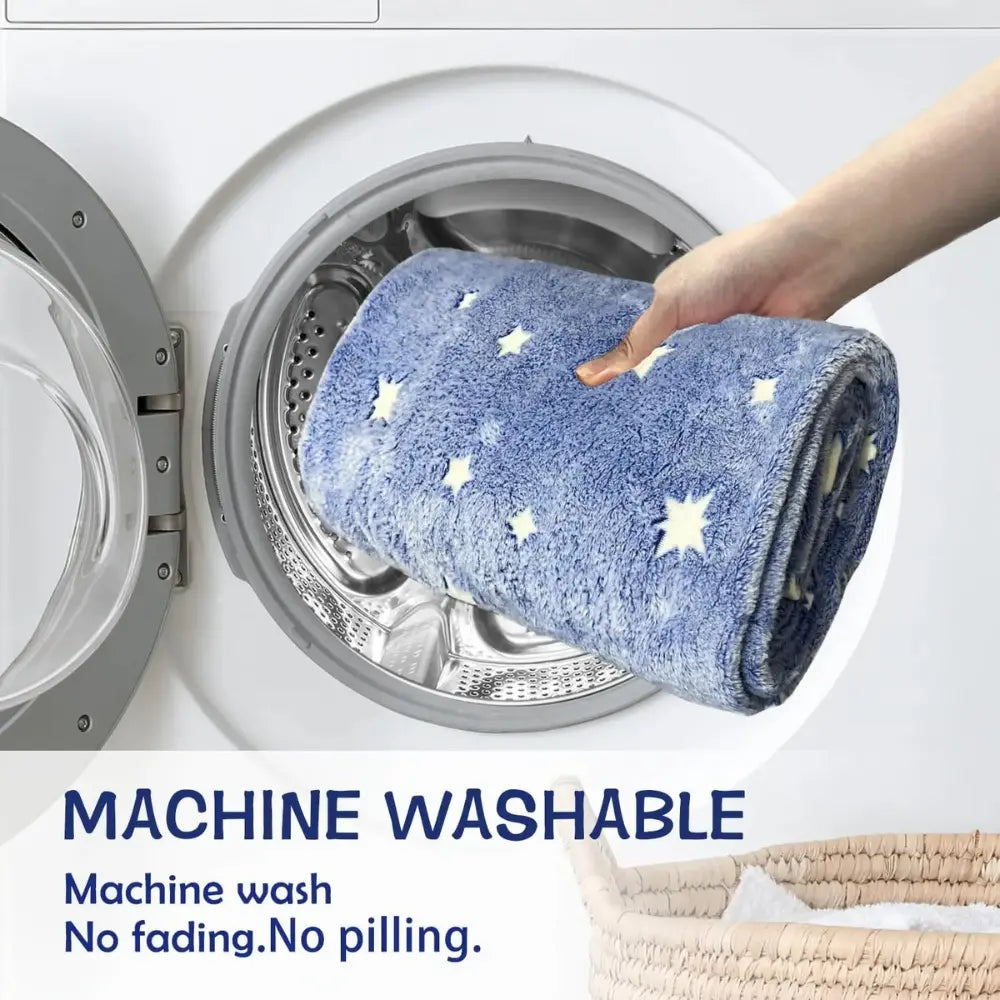 A hand places an ultra-soft 'Glow in the Dark Blanket' by Alaya Junior, featuring blue butterfly and star patterns, into a front-loading washing machine. Text below reads "Machine Washable" and "Machine wash. No fading. No pilling." A wicker basket is partially visible in the bottom right corner.