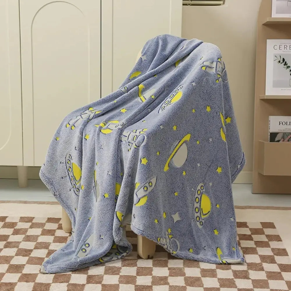 An Alaya Junior Glow in the Dark Blanket, featuring a soft gray fabric adorned with colorful space-themed prints, including yellow and white rocket ships, planets, and stars, is draped over a light-colored chair. In the background are a white cabinet and a bookshelf filled with various folders and items.