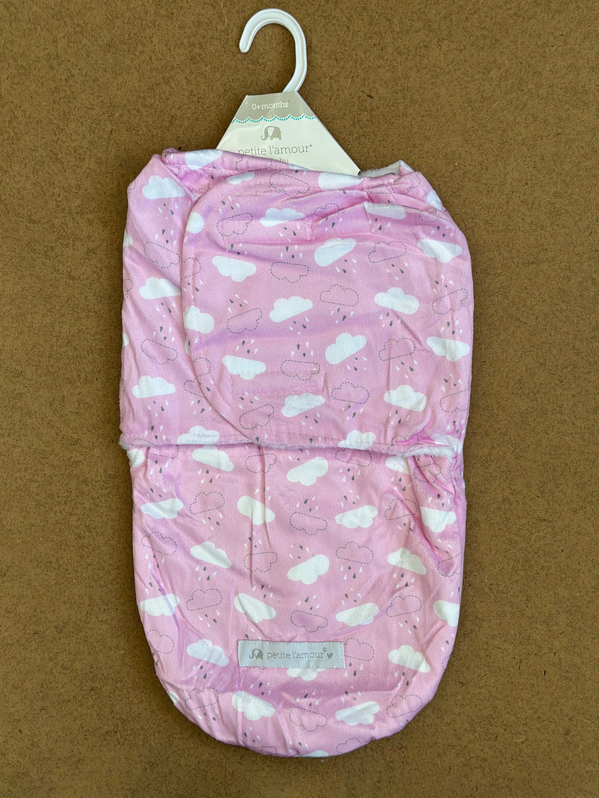 A packaged pink baby swaddle with a cloud pattern from the brand "Alaya Junior" hangs on a white plastic hanger against a brown background. This Adjustable Cloudy Sherpa-Lined Winter Swaddle by Little Sparks features an envelope design, ideal for gently wrapping infants.