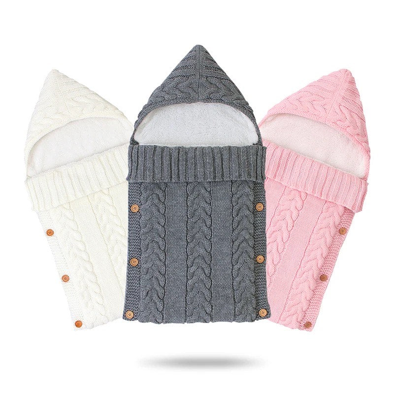 Three Alaya Junior Newborn Baby Sleeping Bags with Sherpa Lining (0-7 Months) are displayed, each featuring a hood and button fastenings. These sleeping bags are arranged side by side, with colors from left to right: white, gray, and pink. They boast a cozy cable knit pattern and adjustable Velcro closures for secure swaddling.