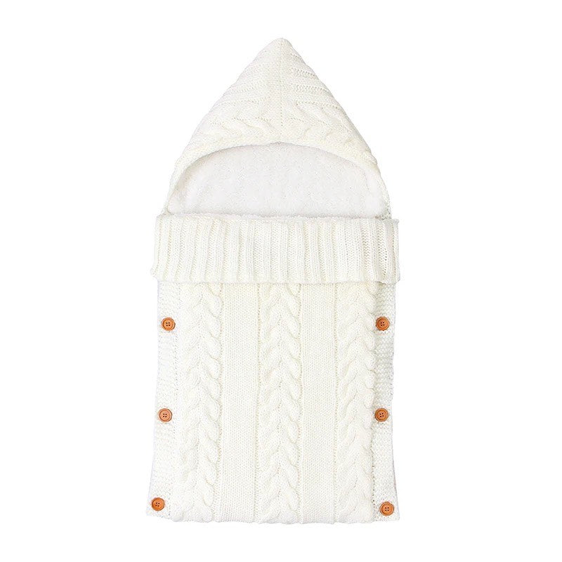 The Alaya Junior Newborn Baby Sleeping Bag with Sherpa Lining (0-7 Months) is an ivory-colored, knitted baby sleeping bag with a hood. It features a cable knit pattern and wooden buttons along the sides for easy closure. The soft, cozy material offers warmth and comfort, ensuring your little one stays snug and secure.