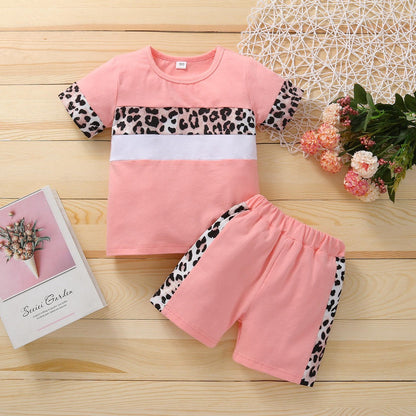 Laid out on a wooden surface is a pink PATPAT Two-piece leopard patchwork outfit for toddler girls. The short-sleeved top and shorts feature pink fabric with white and leopard-print accents. Nearby, a pink book with a floral cover and a small bouquet of flowers rest on a white decorative mat, making it perfect for showcasing infant clothing in Pakistan.