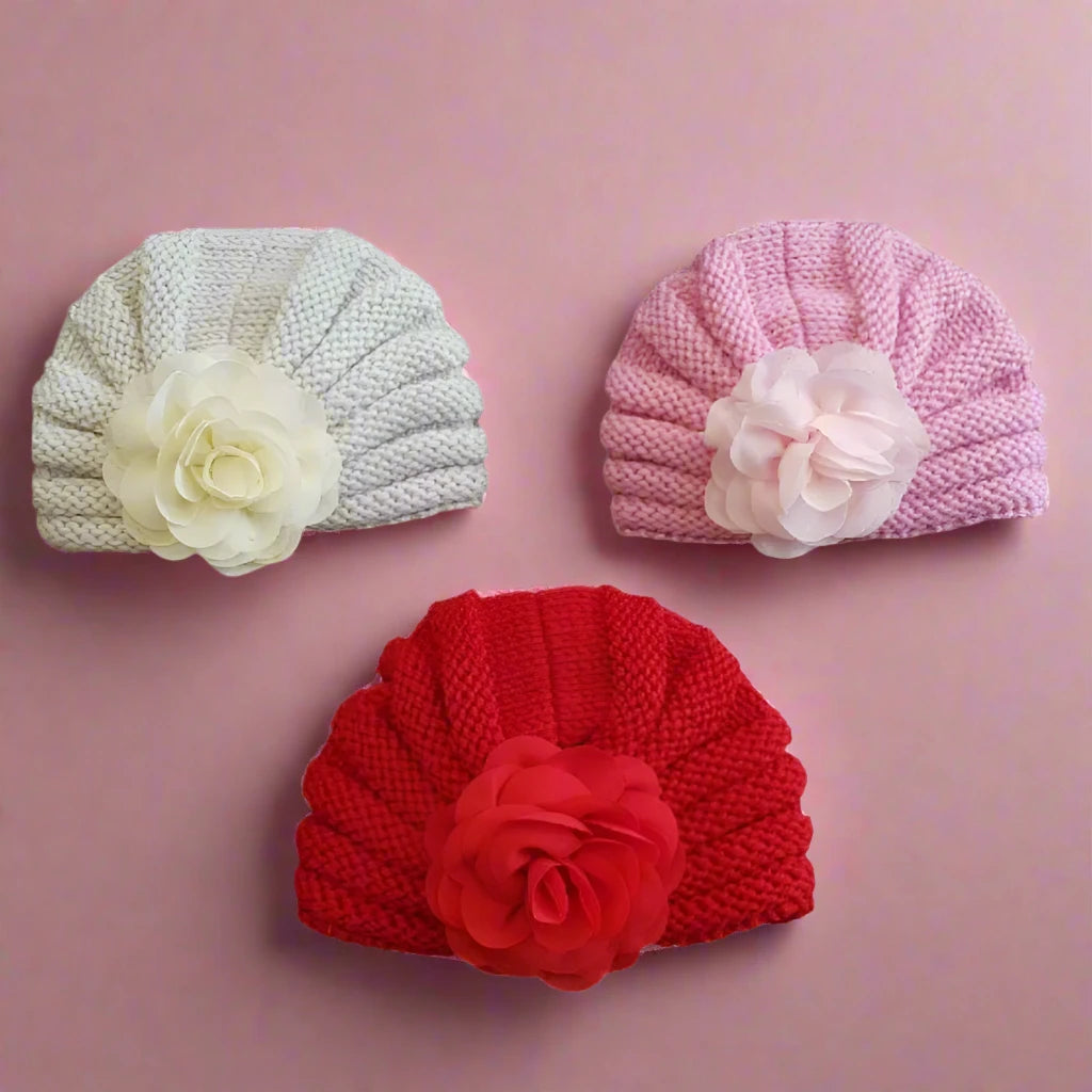 Three soft baby girl caps from Alaya Junior, arranged on a pink background. The top left cap is white with a large white flower, the top right cap is light pink with a light pink flower, and the bottom center cap is red with a red flower. These comfortable designs make an ideal baby gift from the Pack of 3 Soft Baby Girl Caps with Floral Design collection.