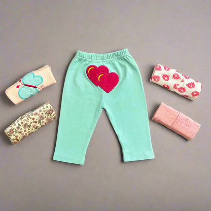 A set of light blue Alaya Junior trousers, featuring heart-shaped patches on the back, is laid flat against a gray background. Surrounding the trousers are five neatly rolled-up baby clothes in various colors and patterns, including florals, strawberries, and polka dots—an ideal collection for newborn and infant clothing in Pakistan.