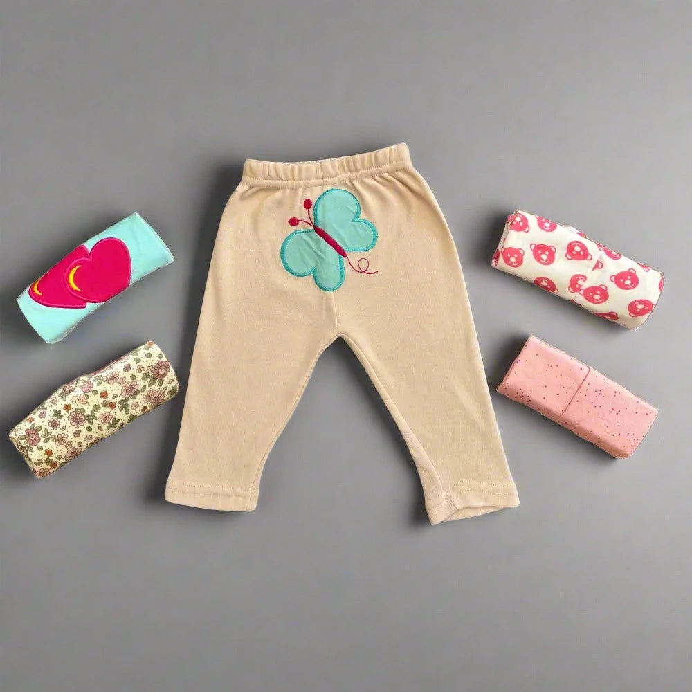 The "Pack of 5 Baby Girl Trousers Soft Fabric" from Alaya Junior features a tan pair of baby pants with a butterfly design in the center, surrounded by four rolled-up baby pants in various patterns: one light blue with a heart, one with strawberries, one with flowers, and one pink with dots. These trousers are perfect examples of the quality infant clothing that Pakistan offers, beautifully showcased against a gray background.