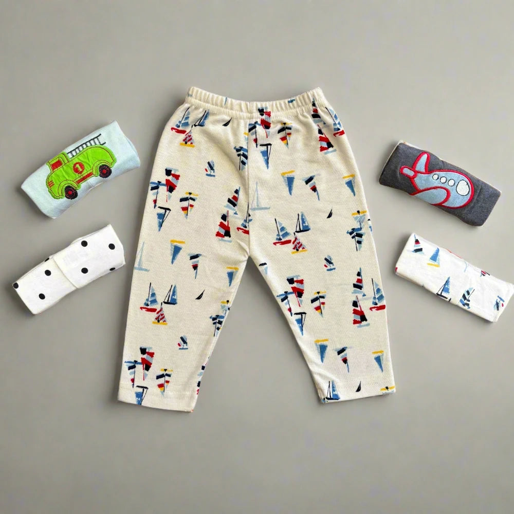 Children's beige pants from Alaya Junior feature colorful sailboat designs, surrounded by folded shirts with vehicle-themed embroidery, including a green vehicle, black dots, and a red airplane. These soft and comfortable unisex baby trousers are presented on a neutral background alongside abstract patterns.