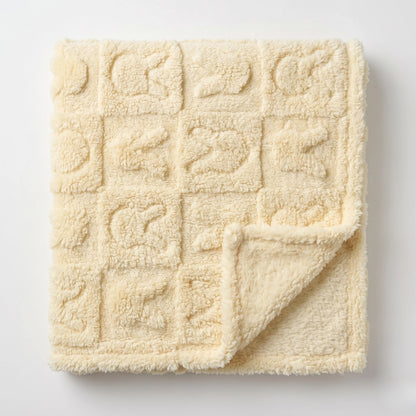 A cream-colored plush blanket with a textured grid pattern of raised square motifs, shown folded with one corner turned over on a light gray background.