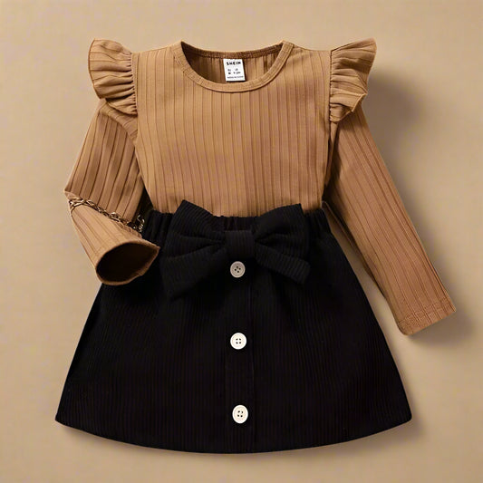 Introducing the SHEIN Baby Girl Ruffle Trim Tee & Bow Front Skirt: a chic ensemble with a ribbed caramel long-sleeve top, ruffled shoulders, and a black bow skirt adorned with white buttons. A chain detail on one sleeve adds flair.