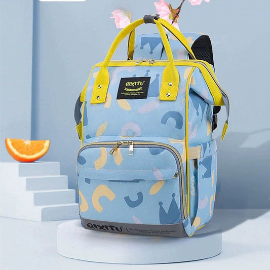 Introducing the Alaya Junior Diaper Bag Backpack: a chic light blue backpack adorned with yellow straps and zippers, featuring eye-catching abstract shapes and crown designs. This practical design includes a front pocket, two side pockets, and a convenient top handle. Pictured on a sleek white platform next to a slice of orange and pink flowers, it's the perfect accessory for all your newborn baby essentials.