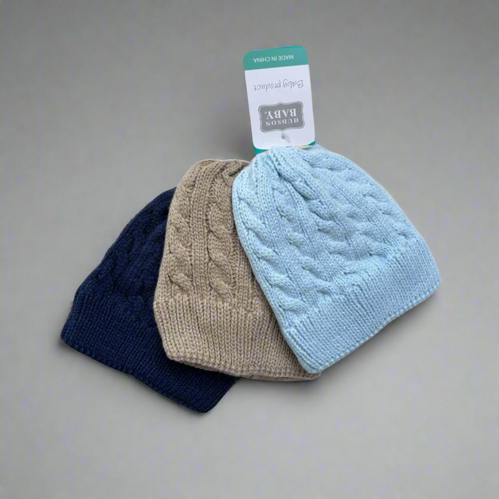 Displayed against a pristine white background with decorative blue accents are the three baby boys' knitted caps from the Pack of 3 Hudson Baby Knitted Caps – Boys (0-12 Months) by Alaya Junior. These caps, suitable for 0-12 months, come in elegant shades of blue, brown, and navy and feature an adorable cable knit pattern along with a "Hudson Baby" tag.