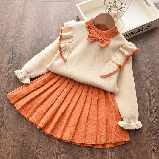 A cozy winter wear ensemble from Alaya Junior is displayed on a wooden surface. The Baby Winter Frock Set for Girls includes a cream-colored ruffle top with rust-orange trim, ruffled sleeves, and a bow on the collar. It is paired with a rust-orange pleated skirt.