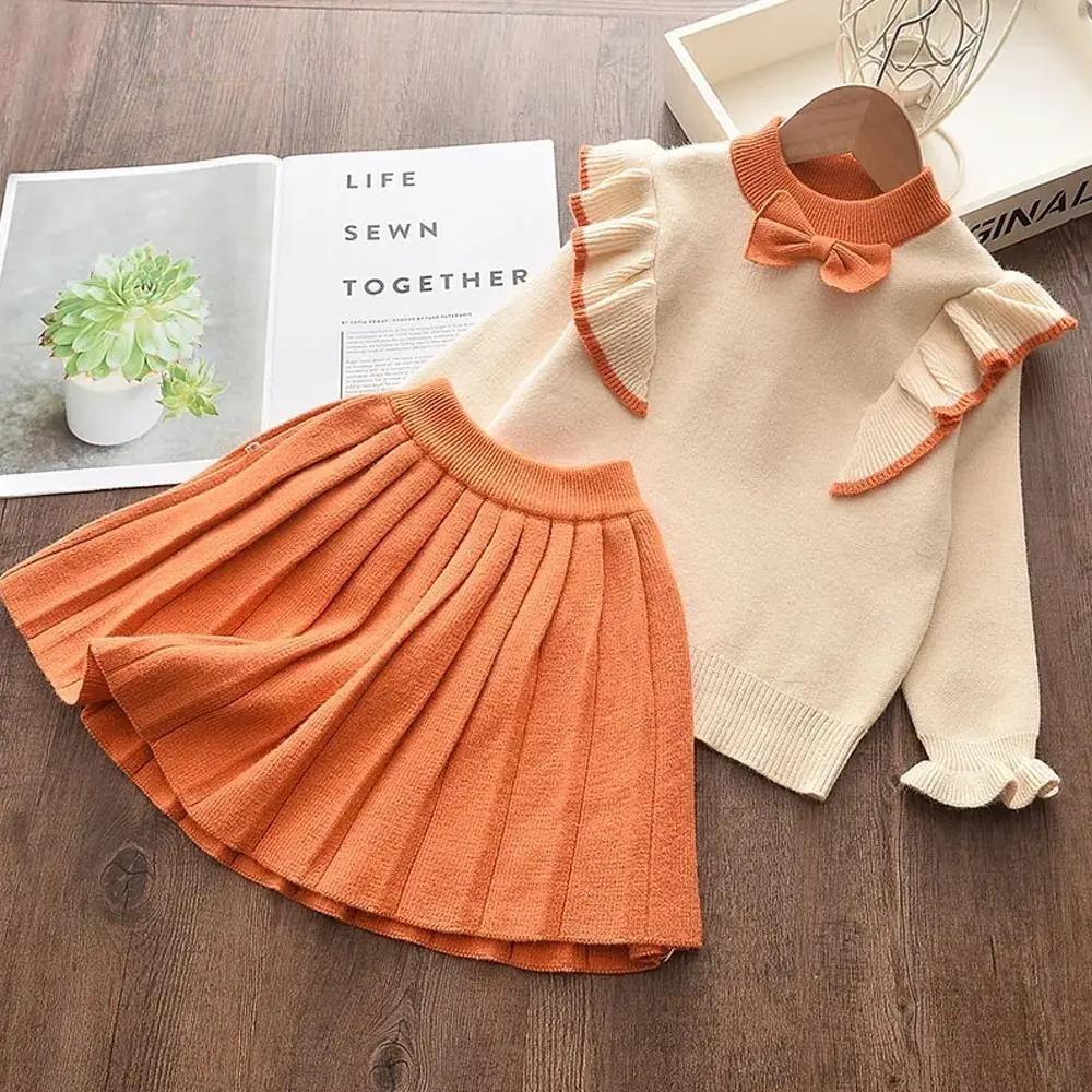 A cozy winter wear ensemble for a girl from Alaya Junior is laid out neatly. This Baby Winter Frock Set for Girls features a creamy white ruffle top with orange details on the shoulders and cuffs, paired with an orange pleated skirt. Beside this charming 2-piece set, an open magazine showcases a succulent plant and text.