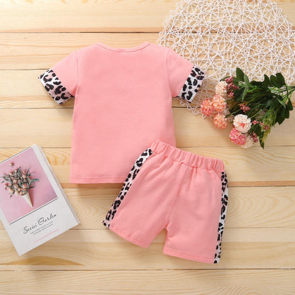 A Two-piece Leopard Patchwork Outfit for Toddler Girls from PATPAT, featuring a pink design with cheetah-print accents on the sleeves and sides of the shorts, is laid out on a wooden surface. Beside the outfit is a decorative white lace mat adorned with a small bouquet of flowers and an open book with a floral cover, making it perfect for showcasing infant clothing from Pakistan.