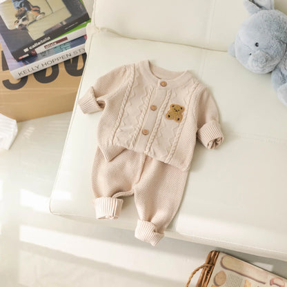 A cozy Baby Knitted 2-Piece Set with Cute Bear Patch from Alaya Junior is laid out on a white leather sofa. The breathable knit fabric of the baby cardigan, featuring an adorable teddy bear patch, is paired with matching pants. Nearby, a blue stuffed animal and a soft toy book are visible, making this ensemble the perfect gift for new parents.