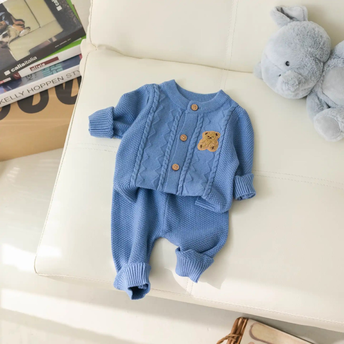 A cozy, blue Baby Knitted 2-Piece Set with Cute Bear Patch from Alaya Junior is laid out on a white couch. The set includes a long-sleeve baby cardigan with wooden buttons and an adorable teddy bear patch, paired with matching pants. A plush gray stuffed bunny sits beside it, making it the perfect gift for new parents. A cardboard box with books is partially visible in the background.