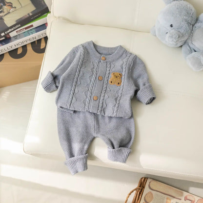 An adorable light gray Baby Knitted 2-Piece Set with Cute Bear Patch from Alaya Junior rests on a white cushioned surface. This breathable knit outfit includes a cable-knit baby cardigan and textured pants, featuring a charming teddy bear applique on the sweater. Nearby, a stuffed gray bear sits alongside some tagged items in the background, making it an ideal gift for new parents.