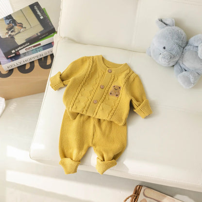 The "Baby Knitted 2-Piece Set with Cute Bear Patch" from Alaya Junior in a cheerful yellow color is displayed on a cream-colored couch. The breathable knit fabric set includes a cardigan adorned with an adorable small bear patch near the chest and matching pants. A gray stuffed bunny sits next to the outfit, making it the perfect gift for new parents. Books and a brown paper bag are visible in the background.
