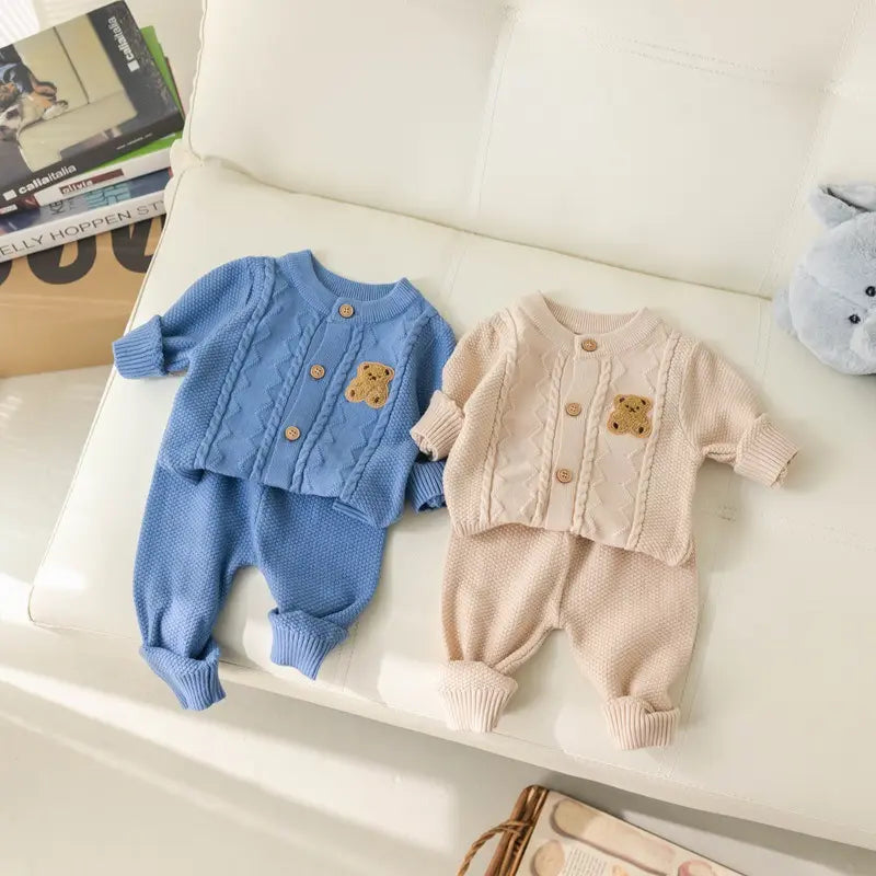 Two baby outfits from the Alaya Junior collection are displayed on a white cushioned seat. Both Baby Knitted 2-Piece Sets with Cute Bear Patch are made from breathable knit fabric and include matching cardigans and pants. One set is blue, and the other is beige, each featuring an adorable teddy bear patch on the baby cardigan. In the background, there's a pile of books and a plush toy.