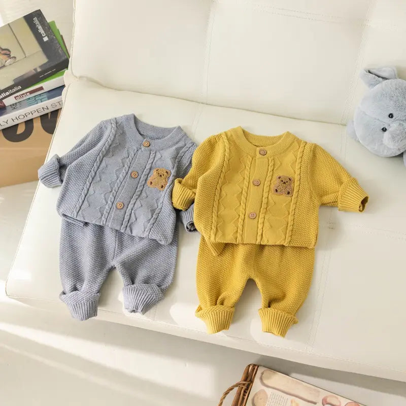 Two baby outfits from Alaya Junior's Baby Knitted 2-Piece Set with Cute Bear Patch lay on a white couch: a gray ribbed knit set with wooden buttons and a small teddy bear patch on the chest, and a yellow ribbed knit set crafted from breathable knit fabric featuring the same design. A blue stuffed toy is visible in the corner, making this scene the perfect gift for new parents.