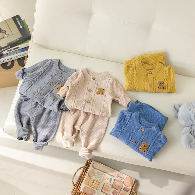 A cozy display of the Alaya Junior Baby Knitted 2-Piece Sets with Cute Bear Patch, featuring four outfits in grey, beige, yellow, and blue. Each set includes a sweater adorned with a teddy bear patch on the chest and matching pants. Made from breathable knit fabric, these charming ensembles are the perfect gift for new parents. Carefully arranged on a white cushioned surface with a plush toy nearby, they make an inviting presentation.