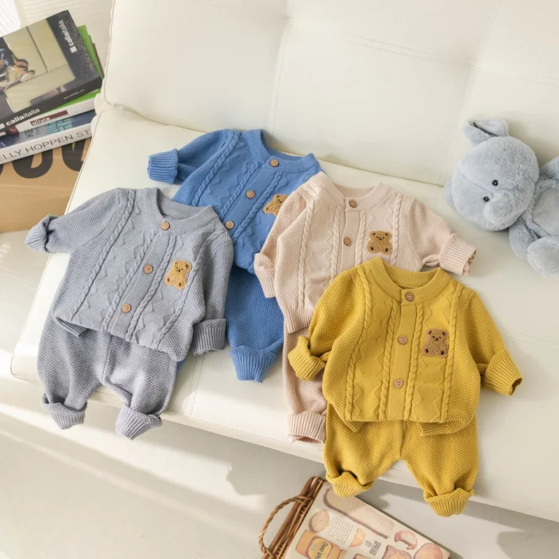 Four sets of the Alaya Junior Baby Knitted 2-Piece Set with Cute Bear Patch are laid out on a white surface. Each outfit includes a baby cardigan with wooden buttons and matching pants, crafted from breathable knit fabric. The outfits come in gray, blue, beige, and yellow, each featuring a small bear applique. Nearby is a plush elephant toy—an ideal gift for new parents.