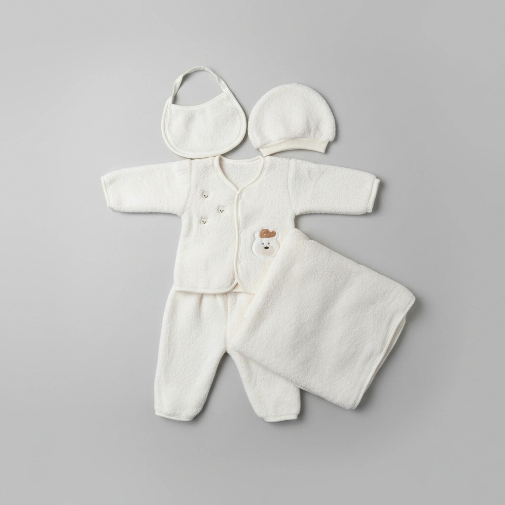 A soft cream-colored baby clothing set laid flat on a white faux-fur surface, including a long-sleeve footed romper, matching hat, bib, and a folded blanket or swaddle. The romper has a small bear face detail at the chest.