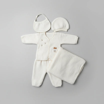 A soft cream-colored baby clothing set laid flat on a white faux-fur surface, including a long-sleeve footed romper, matching hat, bib, and a folded blanket or swaddle. The romper has a small bear face detail at the chest.