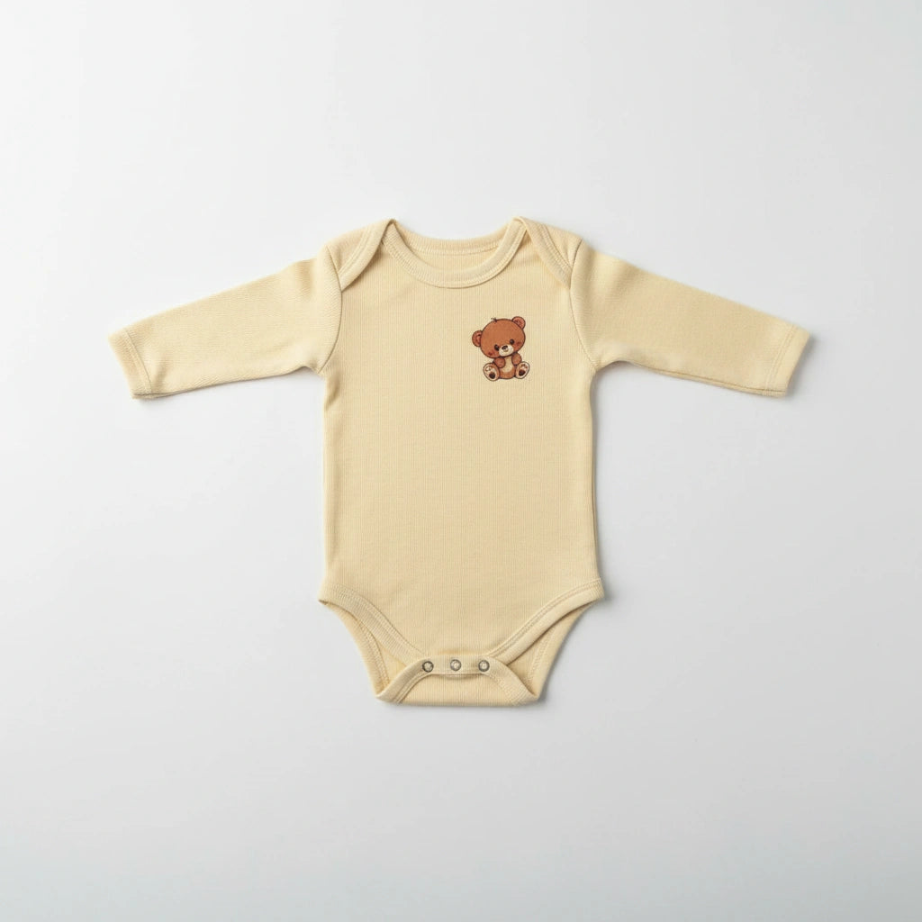 A long-sleeve baby bodysuit in a soft cream-beige color, featuring a small embroidered brown teddy bear on the left chest and three silver snap buttons at the crotch, laid flat on a light gray background.