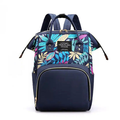 Introducing the Blossom & Bloom Diaper Delight by Alaya Junior - a stylish navy blue backpack adorned with vibrant leaf patterns. The front boasts a large pocket with a gold zipper and an elegant rectangular label that reads "Living Traveling Share." Perfect for carrying baby clothing from Pakistan, this backpack features two padded shoulder straps and convenient side pockets.