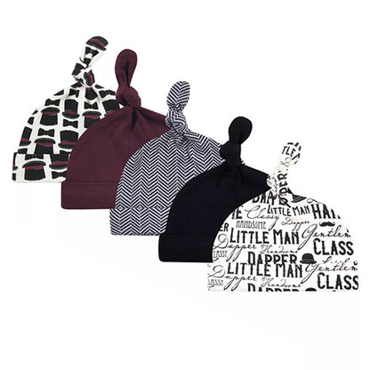 A row of five newborn caps with charming top knots from the Alaya Junior collection features distinct designs: black and white abstract patterns, maroon, a black and white chevron pattern, solid black, and a white cap adorned with printed black words. These Hudson Baby Infant Boy Cotton Caps in the "Vintage Little Man" set are made from ultra-soft cotton to provide comfort for your baby. Suitable for ages 0-6 months.