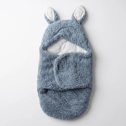 A plush baby swaddle wrap in dusty blue with white inner lining and bunny ears on the hood, shown flat against a light gray background.