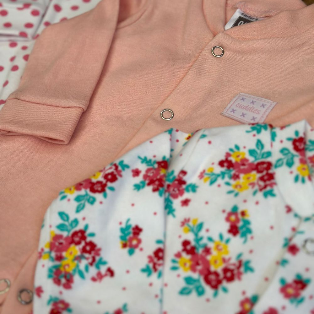 Close-up of newborn baby clothes, including a Carters Pack Of 3 Open Feet Romper (Pink Color) by Alaya Junior with snap buttons on top of a floral-patterned white garment. A grey and black label with stars is visible on the pink item. The background features pink polka dots on white fabric, showcasing adorable infant clothing Pakistan style.