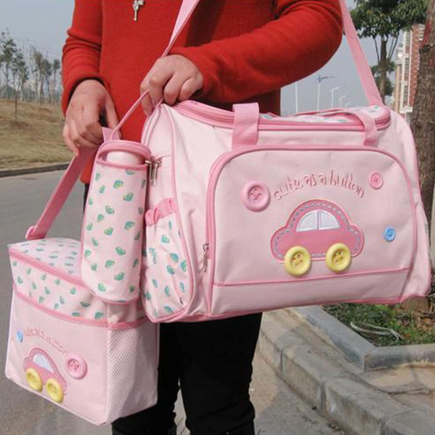 A person in a red top is holding the Alaya Junior Baby Diaper Bag 4 PCS Set, featuring pink bags adorned with car designs. This collection, ideal for baby care essentials, includes a medium-sized tote and a smaller insulated bag complete with bottle holder and lunch bag—crafted from stylish, skin-friendly material highlighted by playful button accents.