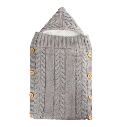 Introducing the Alaya Junior Newborn Baby Sleeping Bag with Sherpa Lining (0-7 Months) – A beautifully knitted baby sleeping bag in soft gray, adorned with a cable knit pattern and featuring a charming pointed hood. Designed for ultimate warmth and comfort, it boasts wooden buttons along the sides for closure and a plush white Sherpa interior lining. Additionally, this cozy baby wrap offers adjustable Velcro closures for easy use. Perfect for keeping your infant snug and secure.