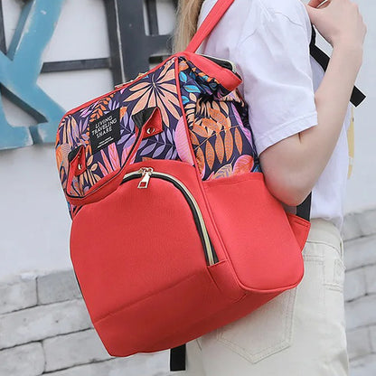 A person wearing a white shirt and light pants is carrying the Blossom & Bloom Diaper Delight backpack by Alaya Junior, featuring a vibrant tropical leaf design with red accents. The backpack includes a front zippered pocket, two buttoned front flaps, and adjustable straps. The background showcases a wall adorned with large letters reminiscent of baby clothing markets in Pakistan.