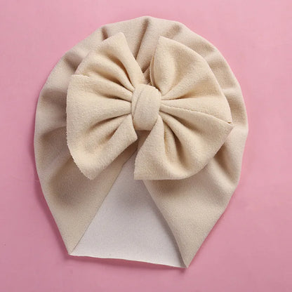 The Alaya Junior PatPat Baby / Toddler Solid Bowknot Hat, featuring a large bow and crafted from soft, textured fabric, graces a pink background. This beige turban-style hat provides a cozy and stylish addition to any little one's wardrobe.