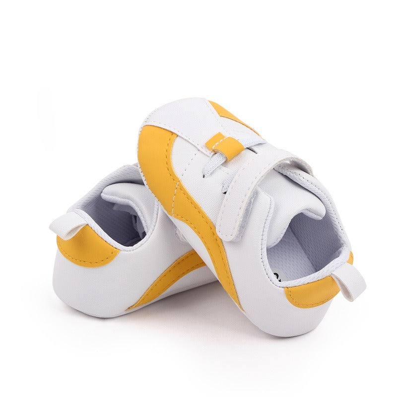 A pair of Alaya Junior's Luxury Baby Shoes for Newborn Girls and Boys in small, showcasing a white and yellow design with Velcro straps. One shoe is standing upright while the other lies on its side, highlighting the simple design and soft PU leather material ideal for newborns. Expertly crafted in Pakistan, both shoes feature a convenient heel tab for easy wearing.