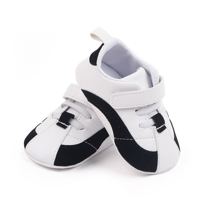 Introducing the Luxury Baby Shoes for Newborn Girls and Boys by Alaya Junior—soft sole anti-slip PU leather sneakers. These white infant shoes, adorned with stylish black accents, make a perfect addition to your newborn's wardrobe. They feature a curved black stripe along the sides and black toes, complemented by white laces and a velcro strap for secure fastening. Ideal for baby clothing in Pakistan, these fashionable sneakers are showcased against a clean, white background.